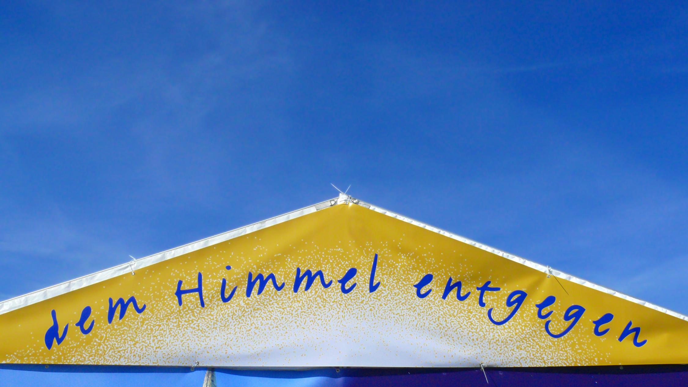 ein goldenes Dreieck mit dem Schriftzug dem Himmel entgegen vor einem blauen Hintergrund