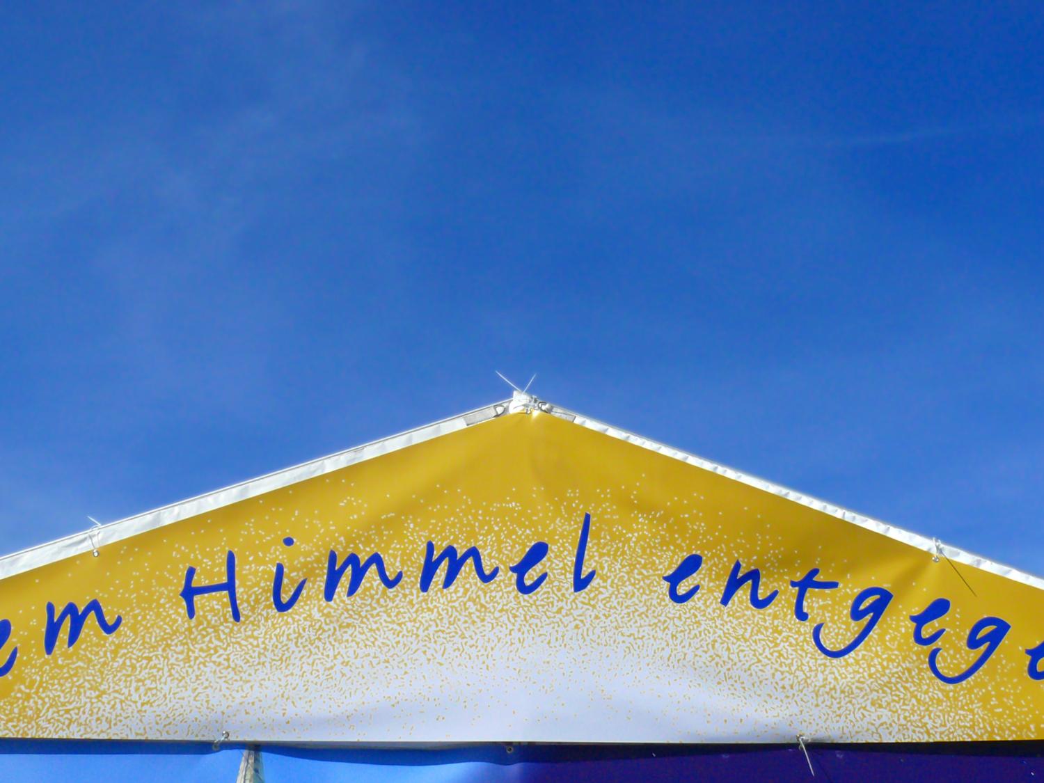 ein goldenes Dreieck mit dem Schriftzug dem Himmel entgegen vor einem blauen Hintergrund