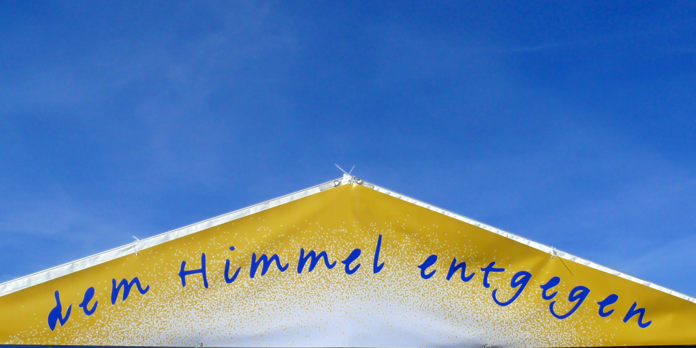 ein goldenes Dreieck mit dem Schriftzug dem Himmel entgegen vor einem blauen Hintergrund