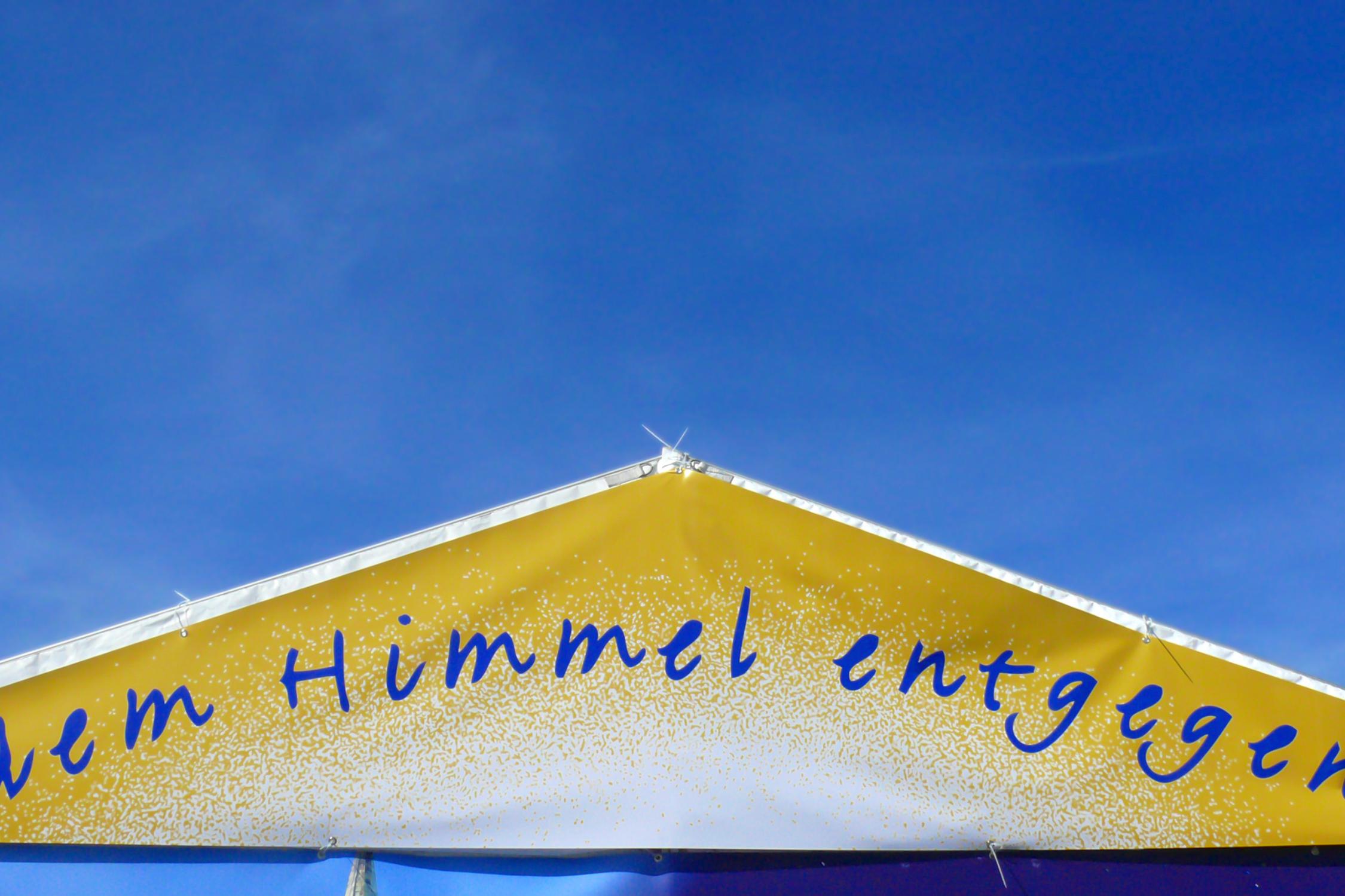 ein goldenes Dreieck mit dem Schriftzug dem Himmel entgegen vor einem blauen Hintergrund