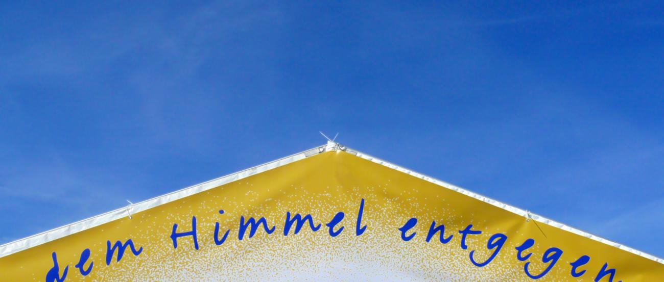 ein goldenes Dreieck mit dem Schriftzug dem Himmel entgegen vor einem blauen Hintergrund