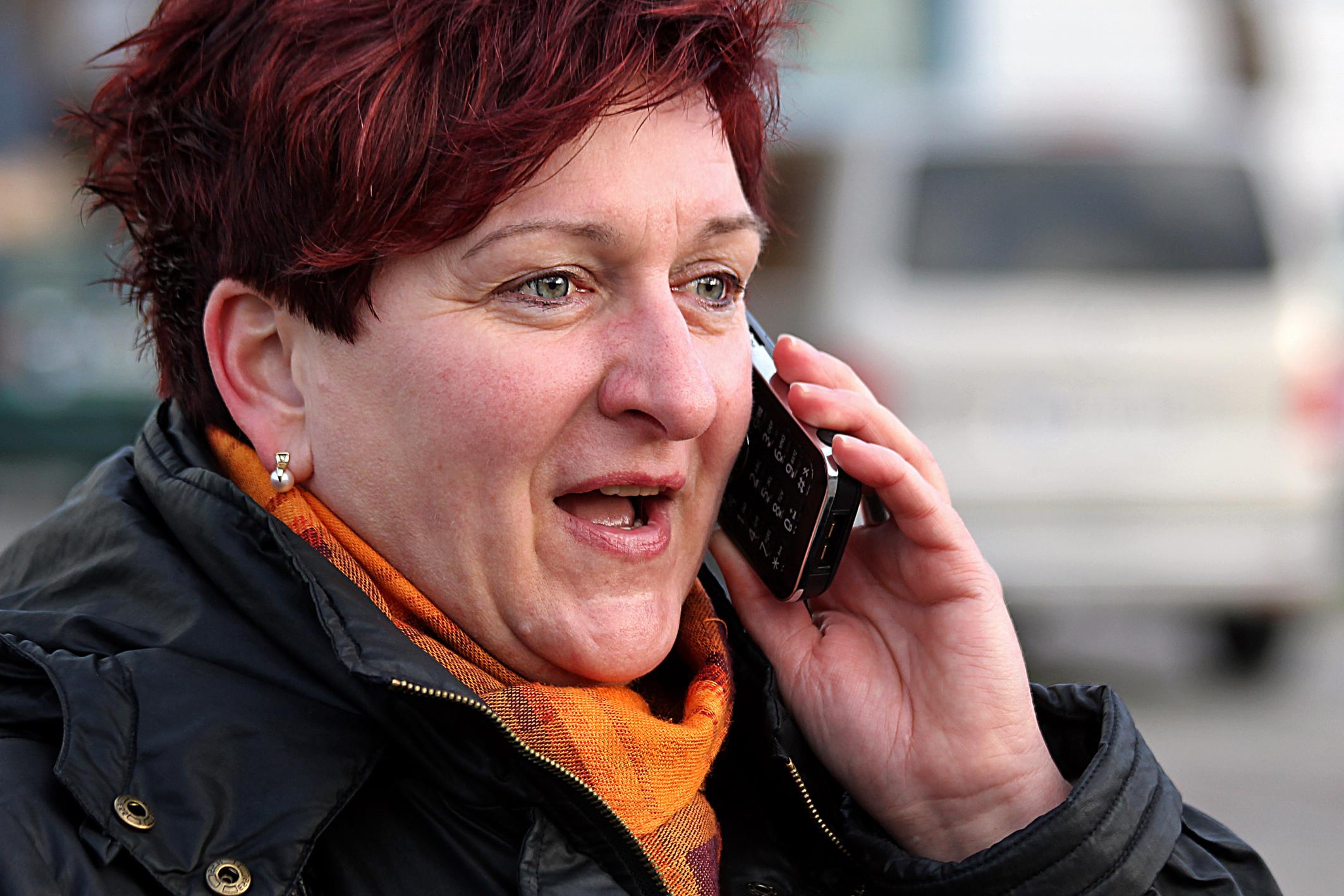 Frau mit rotem Haar telefoniert draußen, trägt Schal und Jacke.