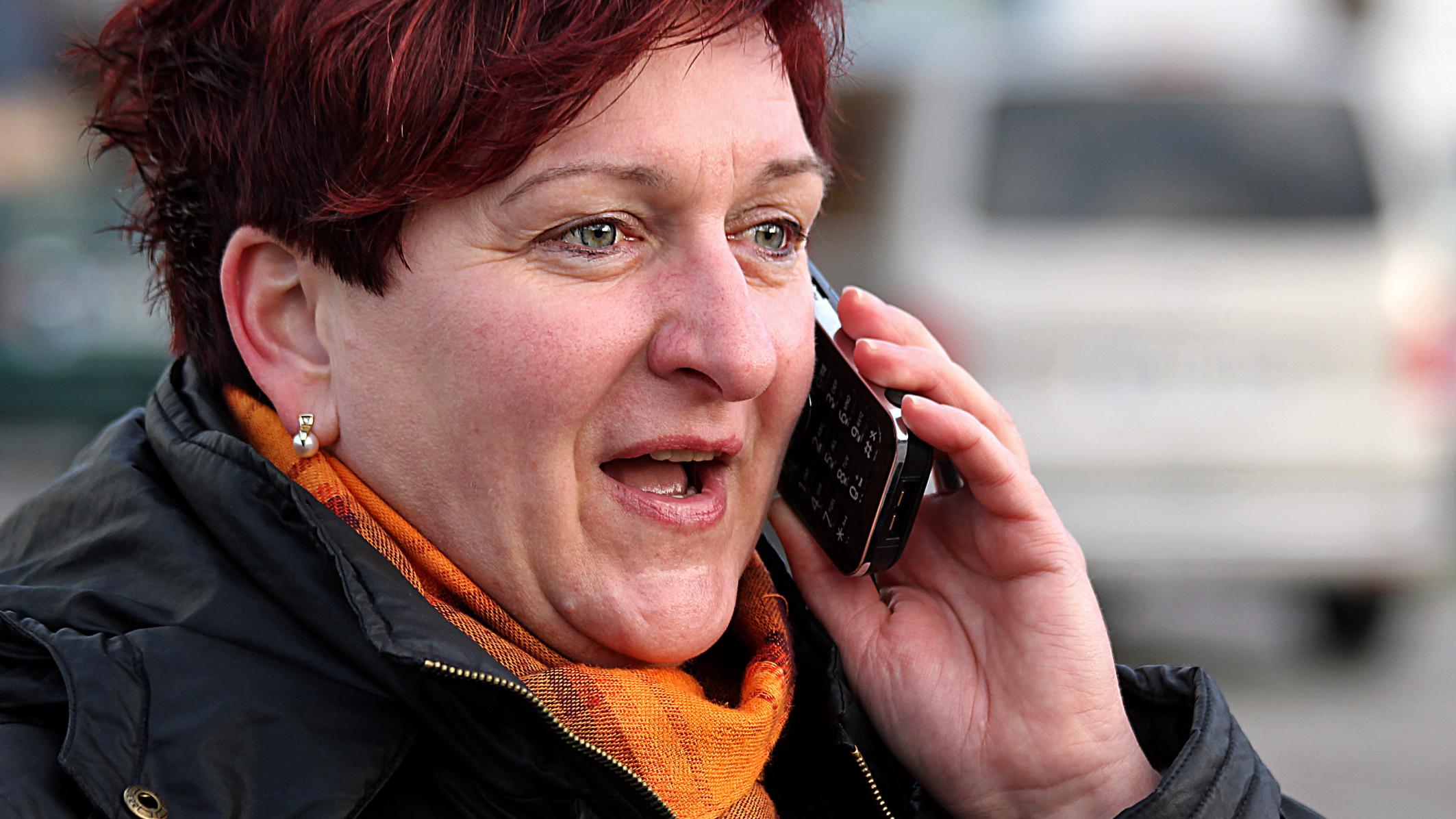 Frau mit rotem Haar telefoniert draußen, trägt Schal und Jacke.