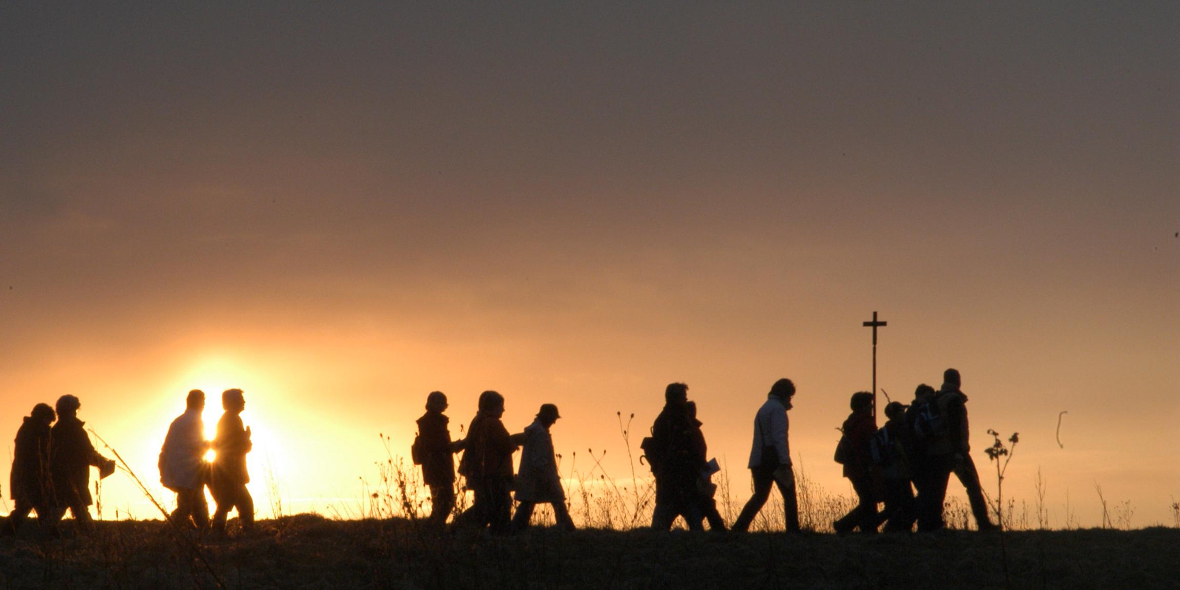 eine Pilgergruppe von 12 Personen zieht über das Bild, im Hintergrund die untergehende Sonne, die Menschen sind als schwarze Silhouetten erkennbar