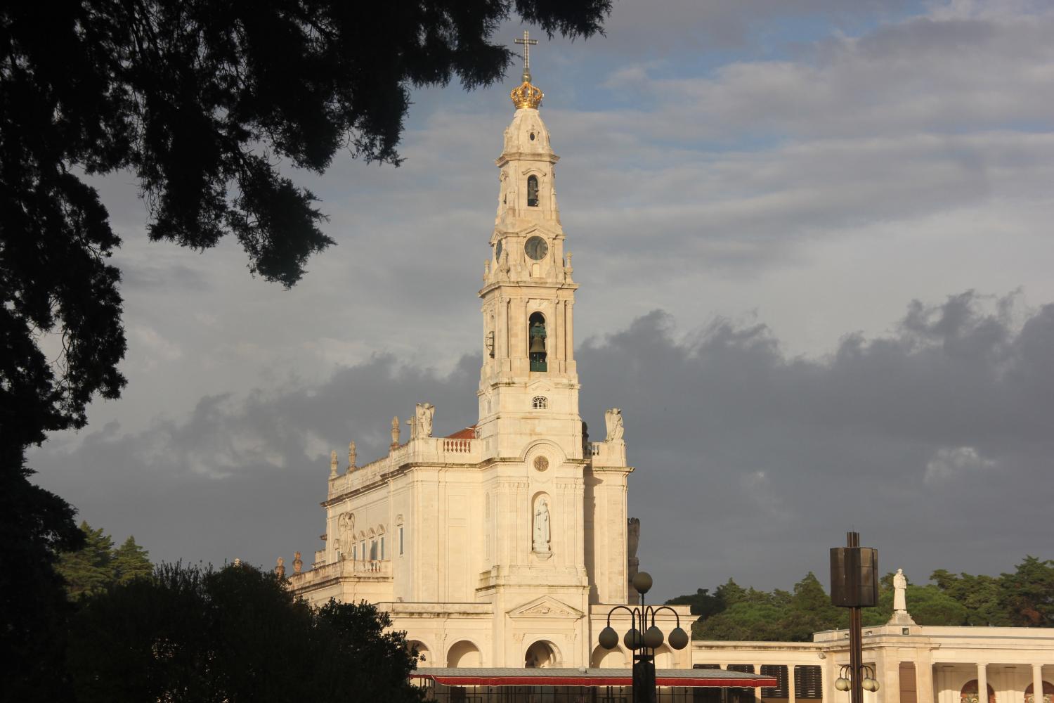 Sonnenbeschienene Basilika unserer lieben Frau des Rosenkranzes in Fátima unter wolkigem Himmel, Bäume im Vordergrund.