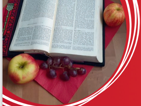 eine aufgeschlagene Bibel auf einer roten Serviette, links unten ein Apfel und einige Trauben, rechts oben neben der Bibel liegt ebenfalls ein Apfel