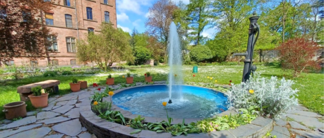 ein Springbrunnen inmitten eines Gartens am linken Rand sieht man das Hauptgebäude des Josefsstift in Trier