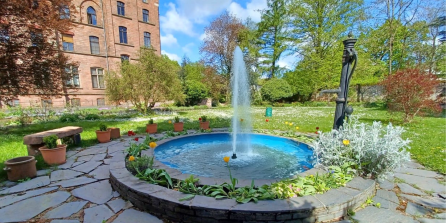 ein Springbrunnen inmitten eines Gartens am linken Rand sieht man das Hauptgebäude des Josefsstift in Trier