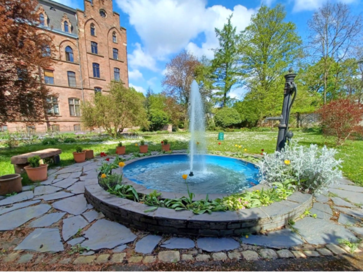 ein Springbrunnen inmitten eines Gartens am linken Rand sieht man das Hauptgebäude des Josefsstift in Trier