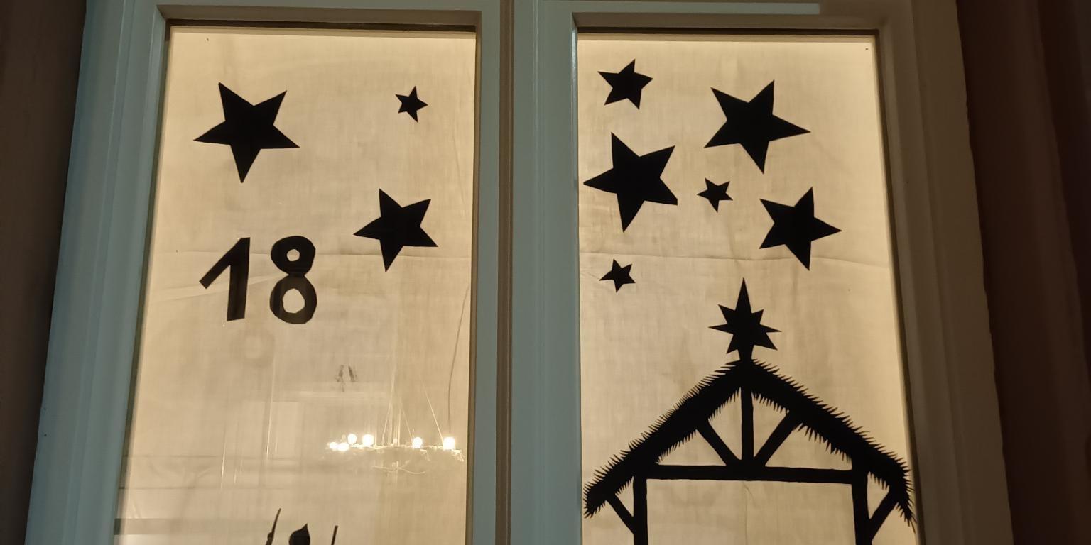 ein Fenster ist mit aus schwarzem Tonpapier ausgeschnittenen Sternen, der Krippe mit Maria, Josef und dem Kind gestaltet. Hinter den Bildern hängt ein weißes Tuch. So sieht es wie eine Scherenzeichnung aus.