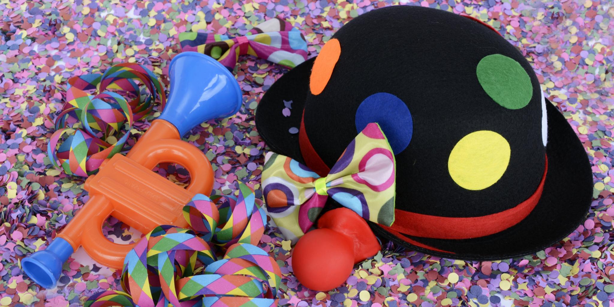 auf dem Bild ist ein schwarzer Clownhut mit bunten Kreisen zu sehen, eine rote Clownnase, eine kleine Tröte und ein paar Luftschlangen