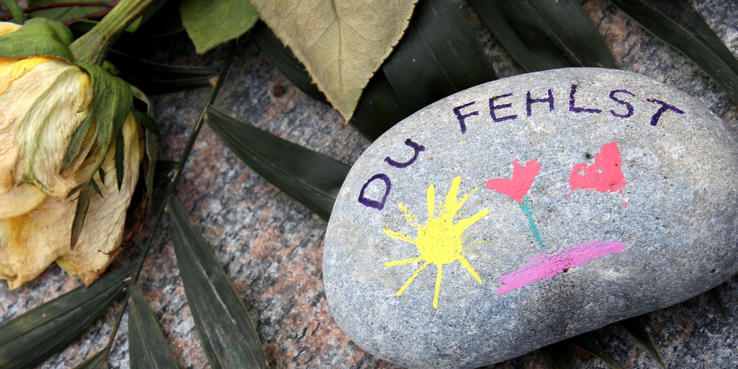 ein mit einer Sonne, Herz und Blume bemalter Stein auf dem Du fehlst geschrieben steht
