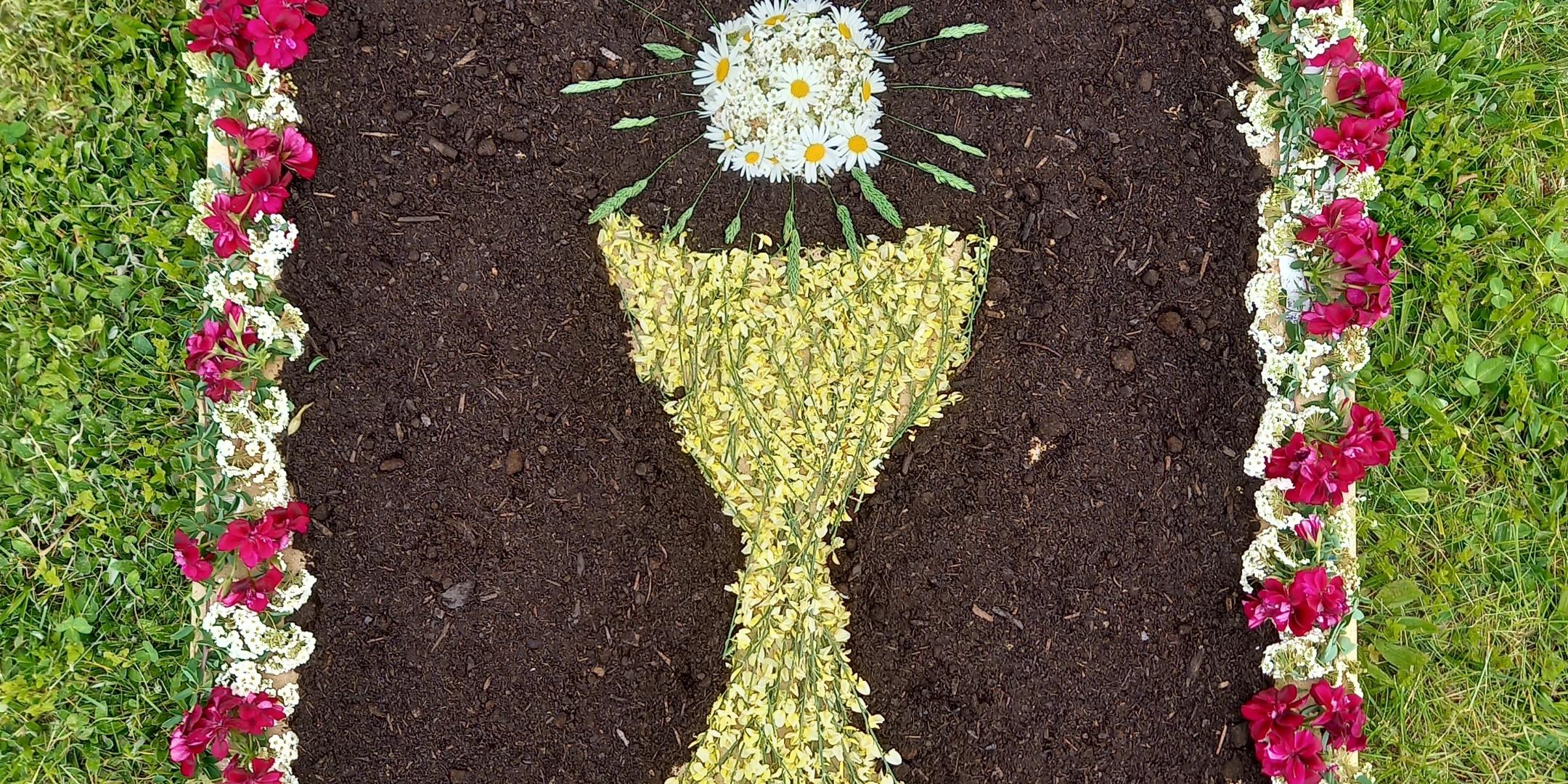 aus Blumen und Fichtennadeln gelegter Teppich abgebildet sind ein Kelch und eine Hostie
