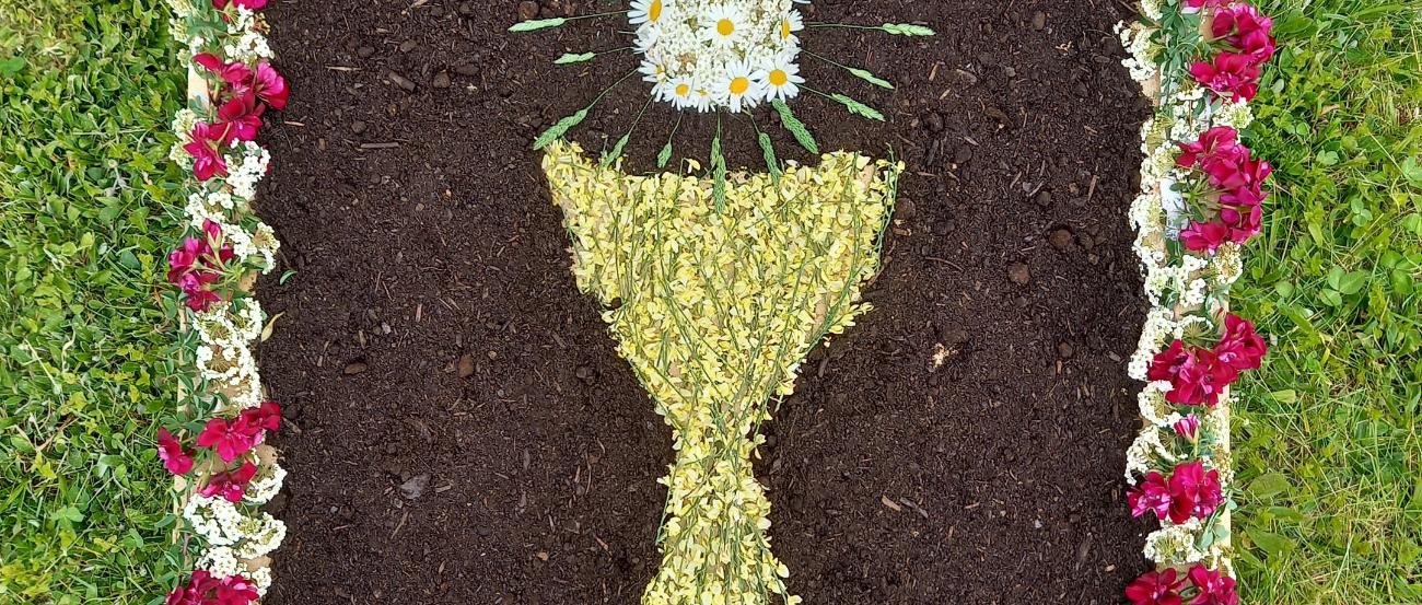 aus Blumen und Fichtennadeln gelegter Teppich abgebildet sind ein Kelch und eine Hostie