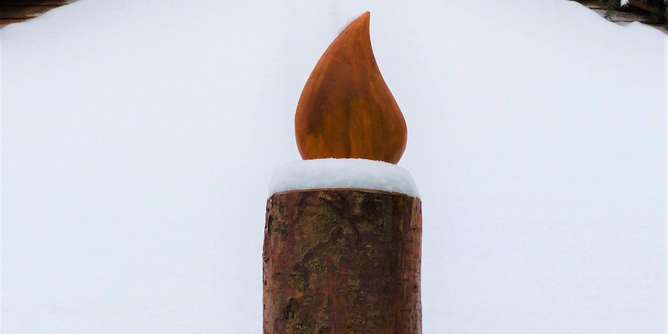 eine große Holzkerze, umgeben von Tannenzweigen auf einem verschneiten Weg