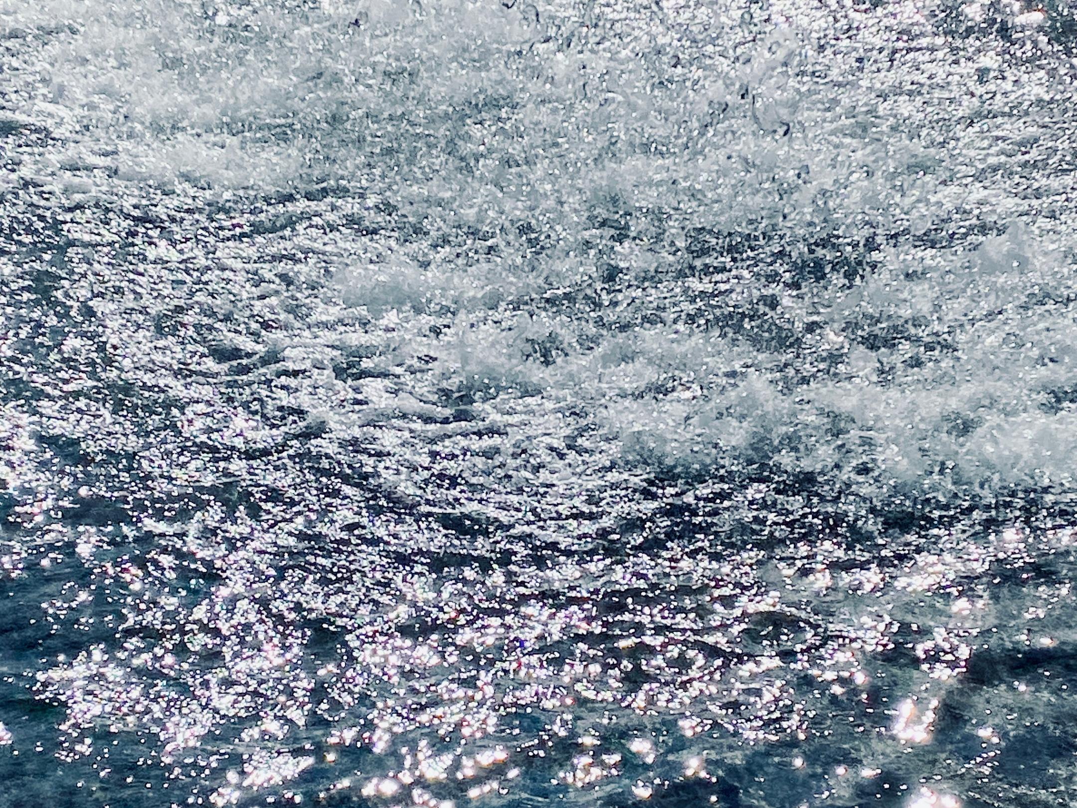 Sprudelnder Springbrunnen in hellem Sonnenlicht. Tausende glitzernde Wassertropfen spritzen über eine unruhige, blau schimmernde Wasseroberfläche, während das Licht in hellen, weißen Reflexionen auf dem Wasser tanzt.
