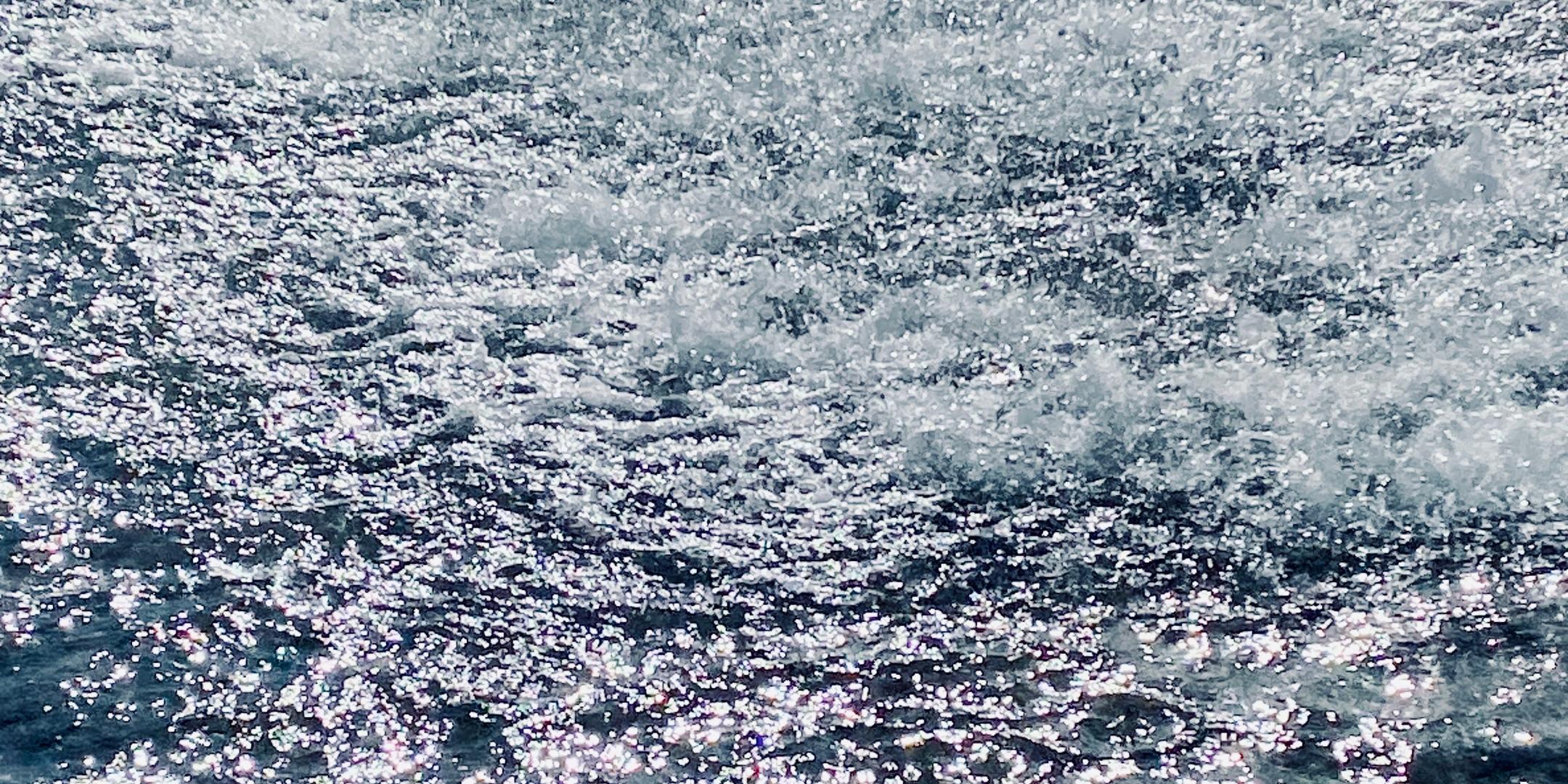 Sprudelnder Springbrunnen in hellem Sonnenlicht. Tausende glitzernde Wassertropfen spritzen über eine unruhige, blau schimmernde Wasseroberfläche, während das Licht in hellen, weißen Reflexionen auf dem Wasser tanzt.
