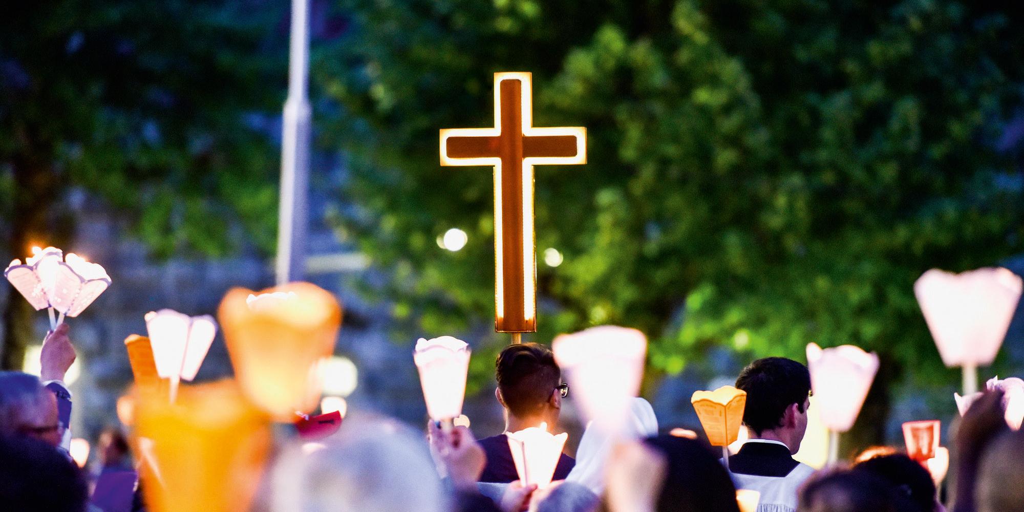 Abendliche Lichterprozession in Lourdes Menschen tragen Kerzenlampions hinter einem roten Vortragekreuz