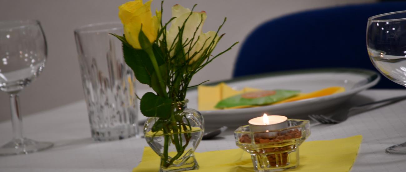 Gelbe Rose in Vase, Teelicht und gedeckter Teller auf weißem Tisch mit gelber Serviette.