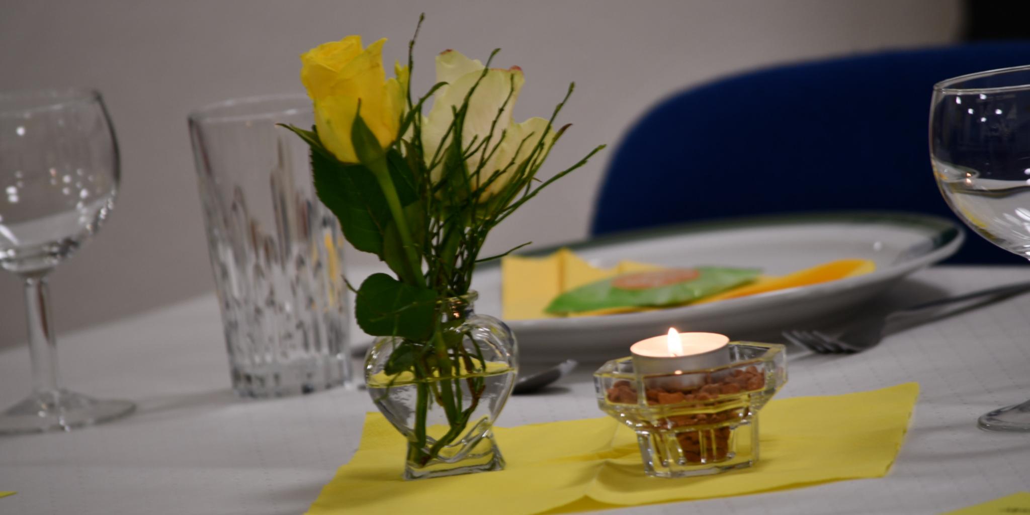 Gelbe Rose in Vase, Teelicht und gedeckter Teller auf weißem Tisch mit gelber Serviette.