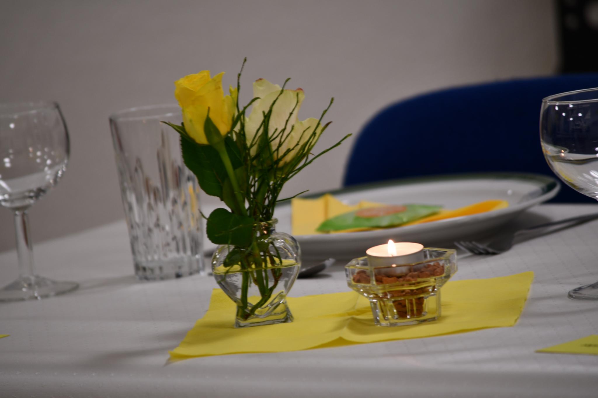 Gelbe Rose in Vase, Teelicht und gedeckter Teller auf weißem Tisch mit gelber Serviette.