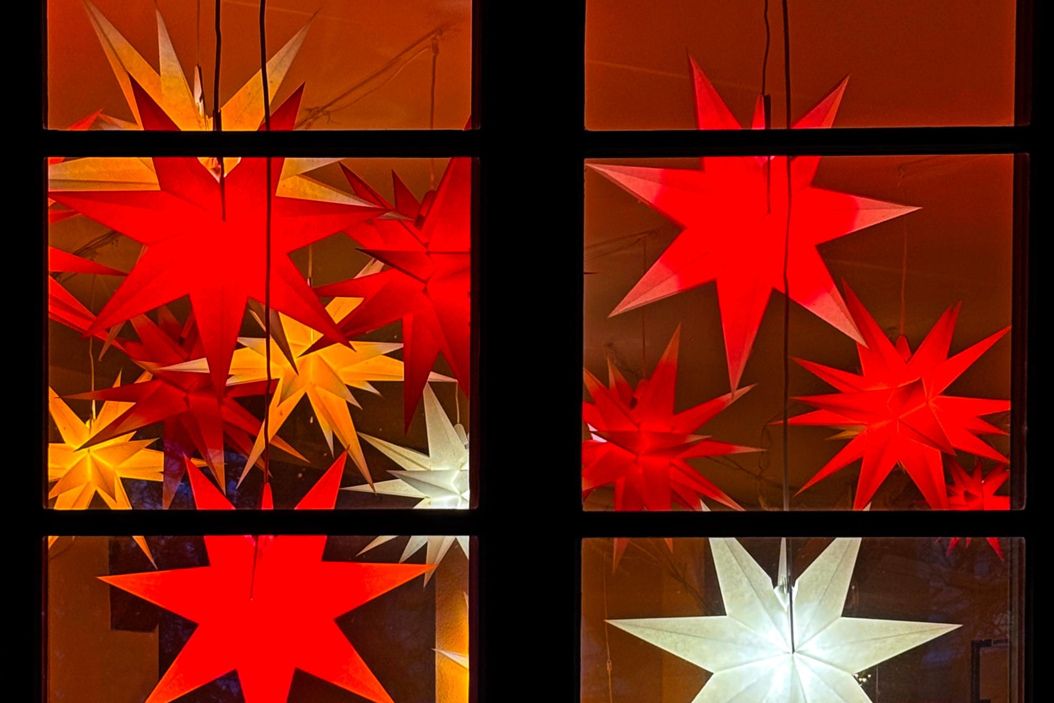 wir schauen auf ein Fenster, dass mit vielen roten, orangen, und 2 weißen Sternen geschmückt ist